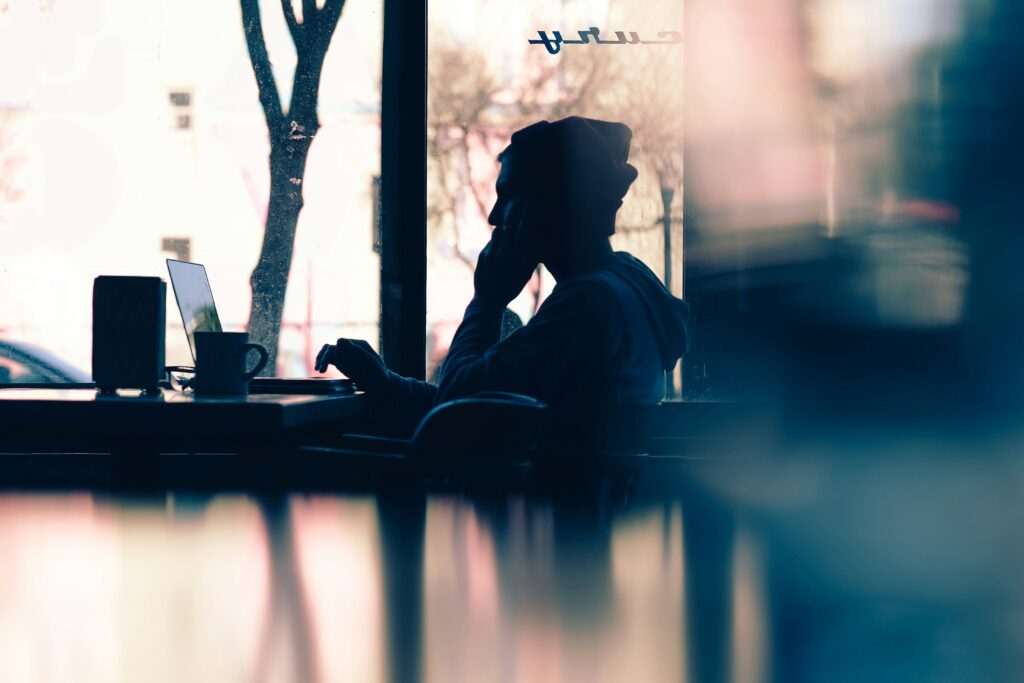 Silhouette von einer Person, die vor einem Fenster sitzt und telefoniert mit Kaffeetasse und Laptop. 