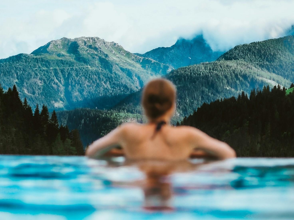 Unscharf: Rücken von Frau in Bikini im Wasser. Scharf: Schöne Ausblick auf Bergspitzen. Laura Chouette / Unsplash.